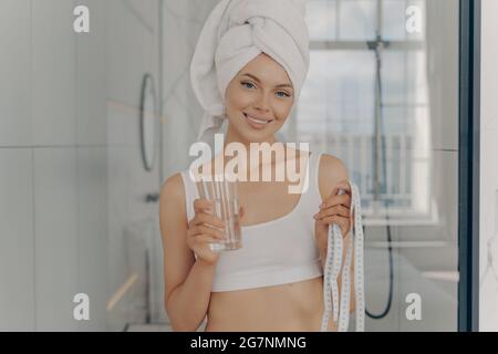 Sorridente giovane donna caucasica attraente con un bicchiere d'acqua in una mano e metro a nastro in un'altra Foto Stock