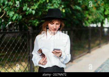 Caucasica ragazza in un cappello tiene gli occhiali e guarda in uno smartphone. Foto Stock