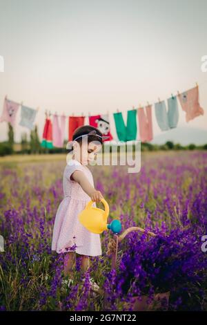 Colpo verticale di ragazza piccola carina che innaffia i fiori di lavanda con attenzione con annaffiatura gialla può Foto Stock