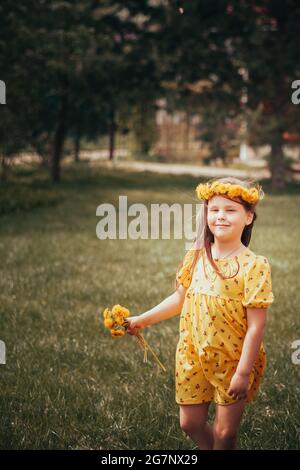 una ragazza con un bouquet di dandelioni e una corona di dandelioni gialli sulla sua testa è in attesa di ospiti di compleanno nel cortile nel giardino a. Foto Stock