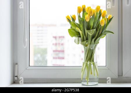 bouquet di tulipani gialli freschi in vaso di vetro sul davanzale della finestra a casa con il paesaggio urbano sullo sfondo Foto Stock