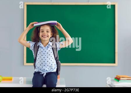 Concetto di ritorno a scuola. La studentessa felice tiene un libro sopra la sua testa ridendo in piedi sullo sfondo di un bordo verde della scuola in classe. Foto Stock