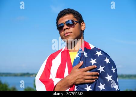 Uomo che guarda la macchina fotografica e tiene orgogliosamente bandiera americana sulle sue spalle Foto Stock