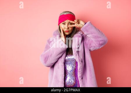 Moda e shopping. Bella donna anziana asiatica in elegante faux pelliccia cappotto e fascia, facendo segno di pace sul viso e guardando sassy e fiducioso a. Foto Stock