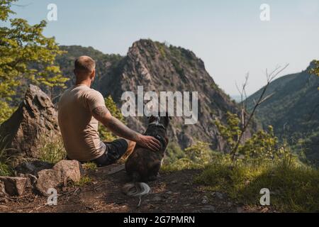 Uomo tedesco adulto seduto su una collina rocciosa con un cane circondato da montagne in Germania Foto Stock