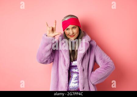 Donna anziana divertente e fredda che mostra rock n roll gesto, guardando sassy, in piedi su sfondo rosa Foto Stock