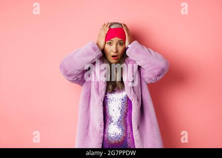 Donna anziana asiatica scioccata e allarmata, indossando un cappotto di pelliccia sintetica viola, tenendo le mani sulla testa e panicking, in piedi angosciati contro sfondo rosa Foto Stock