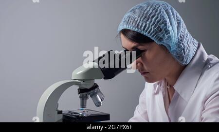 Assistente di laboratorio esperto in cappuccio e cappotto bianco guarda il campione di materiale attraverso il microscopio moderno sul posto di lavoro nello spazio di copia del laboratorio clinico Foto Stock