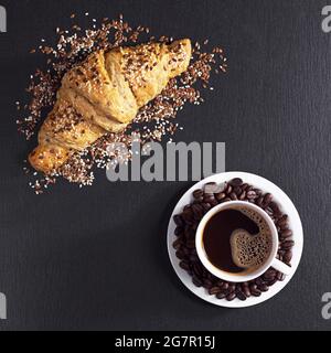 Tazza di caffè e croissant con semi di sesamo per la colazione su fondo di pietra nera, vista dall'alto Foto Stock