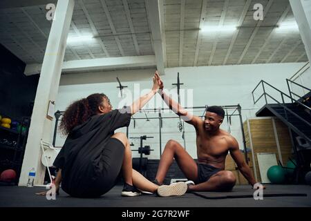 Amici di corsa misti che si adattano a croce in palestra. Uomo personal trainer che dà afroamericano femmina un high-five dopo l'esercizio. Foto di alta qualità Foto Stock