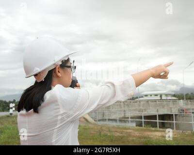 Ingegnere femminile in un casco bianco che utilizza la comunicazione radio con il lavoratore di costruzione presso il cantiere della diga per generare elettricità. Foto Stock