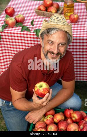 Carismatico agricoltore maturo che tiene la mela rossa e guardando la fotocamera. Concetto di cibo sano. Coltivatore che raccoglie le mele in un frutteto. Foto Stock