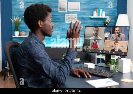 L'uomo afro-americano che parla di videochiamata online che sventola ai colleghi utilizzando la comunicazione con la webcam. Persona nera che lavora da casa mantenendo la distanza mentre utilizza la tecnologia digitale Foto Stock