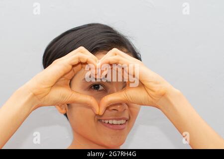 Una giovane donna, capelli corti, bella, sta facendo un segno d'amore con il dito, vicino ai suoi occhi Foto Stock