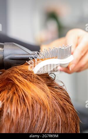 Closeup della mano del padrone con l'aria-asciugando e la spazzola che soffia i capelli rossi femminili in un salone Foto Stock