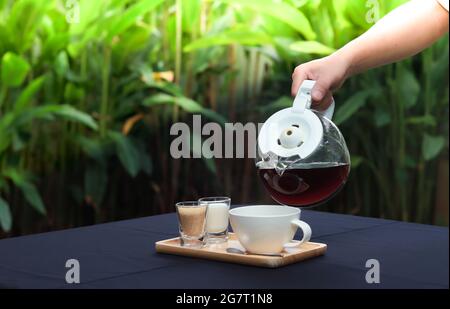 Tenere a mano una pentola di caffè, versando in un bicchiere su un tavolo con un vassoio di legno, latte e zucchero. Atmosfera in un giardino ristorante. Foto Stock