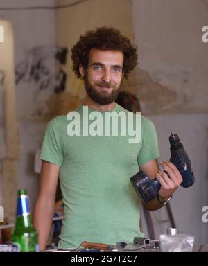 Giovane uomo di riparazione sorridente portandogli in piedi al tavolo da lavoro con un trapano in una mano presso il suo negozio di riparazione bici. 23 luglio 2019. Kiev, Ucraina Foto Stock