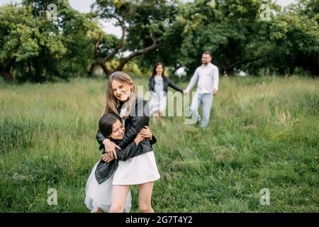 Due allegre sorelle che abbracciano e sorridono insieme nel giardino verde mentre i loro genitori tengono le mani e camminano sullo sfondo. La famiglia felice di quattro persone si godendo del tempo libero all'aria aperta. Foto Stock