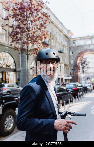 uomo d'affari gioioso e maturo in scooter elettrico blazer e casco Foto Stock