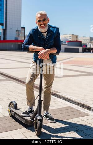 un uomo allegro in occhiali da sole in piedi vicino a e-scooter nella città urbana Foto Stock