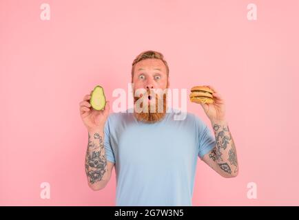 L'uomo stupito con barba e tatuaggi è indeciso se mangiare un avocado o un hamburger Foto Stock