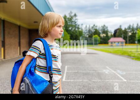 Bel bambino felice vicino alla scuola edificio presso il cortile. Piccolo studente sorridente che porta con sé uno zaino blu. Foto Stock