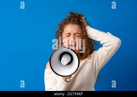 Giovane sorridente espressiva donna caucasica gridò in megafono su sfondo blu Foto Stock