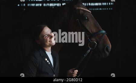 Horsewoman in posa con il suo cavallo marrone sigillo. Sorridendo e tenendo la sua briglia. La ragazza è vestita con un cappotto nero. Il cavallo sta portando i monili della testa. A cavallo. Legame tra il cavallo e il suo padrone. Foto Stock