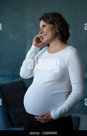 Donna incinta che parla sul telefono tiene lo smartphone, toccandola la pancia, consulta con qualcuno Foto Stock