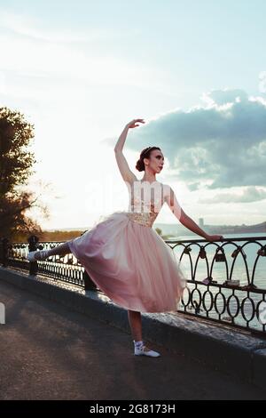 Una giovane ballerina in un abito di seta rosa balla ed esegue esercizi, tenendo su una recinzione in ghisa sul lungofiume del fiume Volga al tramonto Foto Stock
