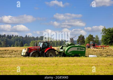 Trattori Massey-Ferguson nel campo del fieno con legatore per rotopressa McHale e rastrello rotante che raccoglie fieno secco in estate. Raasepori, Finlandia. 25 giugno 2021. Foto Stock