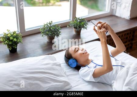 Una bambina di dieci anni ascolta la musica a letto. Ascolta la musica tramite le cuffie e un'applicazione online sul suo smartphone. Cultura giovanile e mo Foto Stock