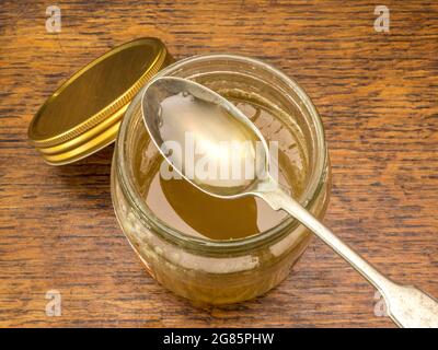 Vista dall'alto di un cucchiaio di miele d'api puro spesso appoggiato sulla parte superiore di un vaso di prodotto aperto, in piedi su un tavolo di legno. Foto Stock