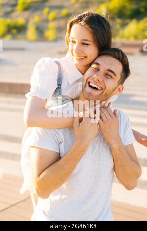Ritratto verticale di giovane coppia allegra che si diverte nel parco cittadino in estate giorno di sole, giovane donna appesa sulle spalle di amante ridente. Foto Stock