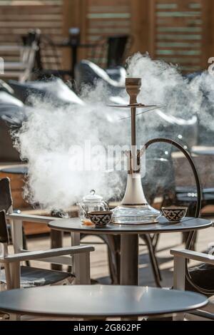 Fumare hookah trasparente sul tavolo sulla terrazza estiva nella lounge hookah fare fumetti nuvole e bere tè Foto Stock