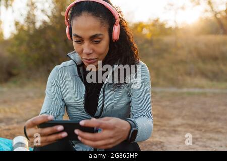 Donna sportiva afro-americana focalizzata in cuffia con cellulare mentre si siede in parco Foto Stock