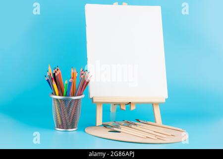 Cavalletto in legno con tela bianca, pennelli e matite colorate su sfondo blu. Foto Stock