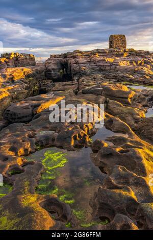 La calda luce del mattino cattura le rocce che circondano la Powder House a Seahouses sulla costa del Northumberland, Inghilterra, Regno Unito Foto Stock