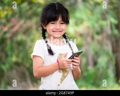 Una ragazza asiatica di sette anni sorride felicemente mentre usa il suo telefono cellulare per cercare informazioni durante un viaggio con la famiglia al parco. Foto Stock