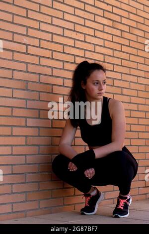 Giovane ragazza sportiva spagnola in posizione squatting dopo un duro allenamento contro un muro di mattoni Foto Stock