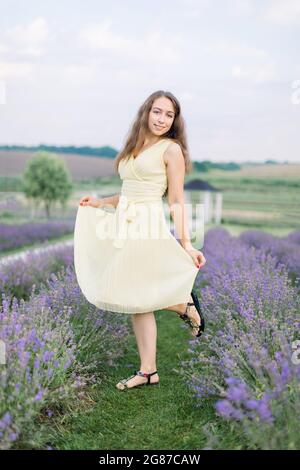 Bella ragazza sul campo di lavanda. Bella donna dai capelli lunghi in un vestito pastello chiaro che posa a macchina fotografica con sorriso nel campo di lavanda al tramonto. Messa a fuoco morbida Foto Stock