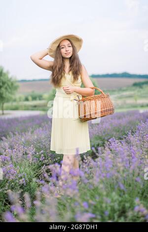 Ritratto a lunghezza intera di una bella giovane donna, indossando un abito beige giallo chiaro, cappello di paglia, camminando con borsa di vimini in estate fiorendo campo di lavanda, godendo profumo di lavanda Foto Stock