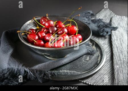 ciliegie rosse fresche in una ciotola grigia con tovaglioli su sfondo scuro, snack estivo sano, spazio copia, messa a fuoco selezionata, profondità di campo stretta Foto Stock