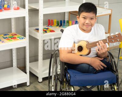 Ritratto di bambino disabile in una sedia a rotelle sta giocando l'ukulele in attività sensoriale. Concetto di apprendimento dei bambini disabili a scuola. Foto Stock