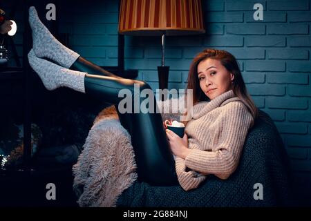 Bella donna con tazza di caffè seduta sulla sedia. Atmosfera di Natale e Capodanno. Tazza di cioccolata calda con marshmallows. Bella ragazza in maglione. Parete grigia in mattoni su sfondo. Vacanza invernale. Foto Stock