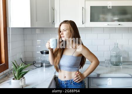 Giovane donna felice che beve caffè o tè sulla cucina scandinava bianca all'interno del Morning.Wearing jeans e crop top. Foto Stock