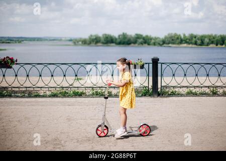 ritratto di stile di vita, il momento del movimento di una ragazza su uno scooter, una ragazza in un vestito guida uno scooter lungo il argine del fiume con una recinzione in ghisa Foto Stock