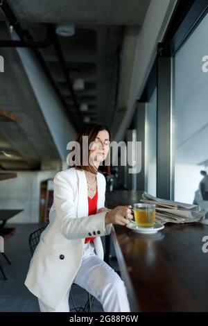 Giovane donna di bellezza millennial rilassarsi al caffè o tè freno in caffè. Indossa una giacca da ufficio bianca e una tazza con tè verde. Giornali su t Foto Stock