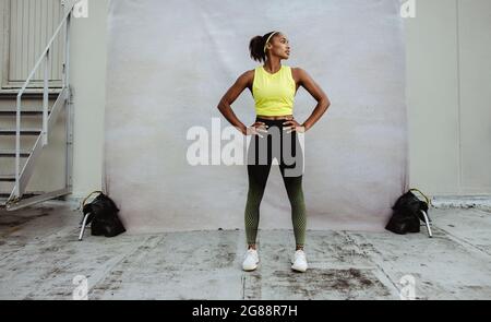 Intera lunghezza di donna africana in abbigliamento sportivo su una terrazza dell'edificio. Atleta donna con corpo in vestibilità in piedi con le mani in vita guardando lontano. Foto Stock