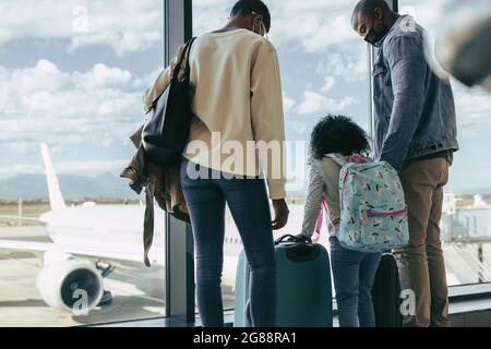 Vista posteriore della famiglia africana in piedi vicino al finestrino del terminal dell'aeroporto con bagagli. Famiglia turistica che aspetta all'aeroporto con i bagagli. Foto Stock